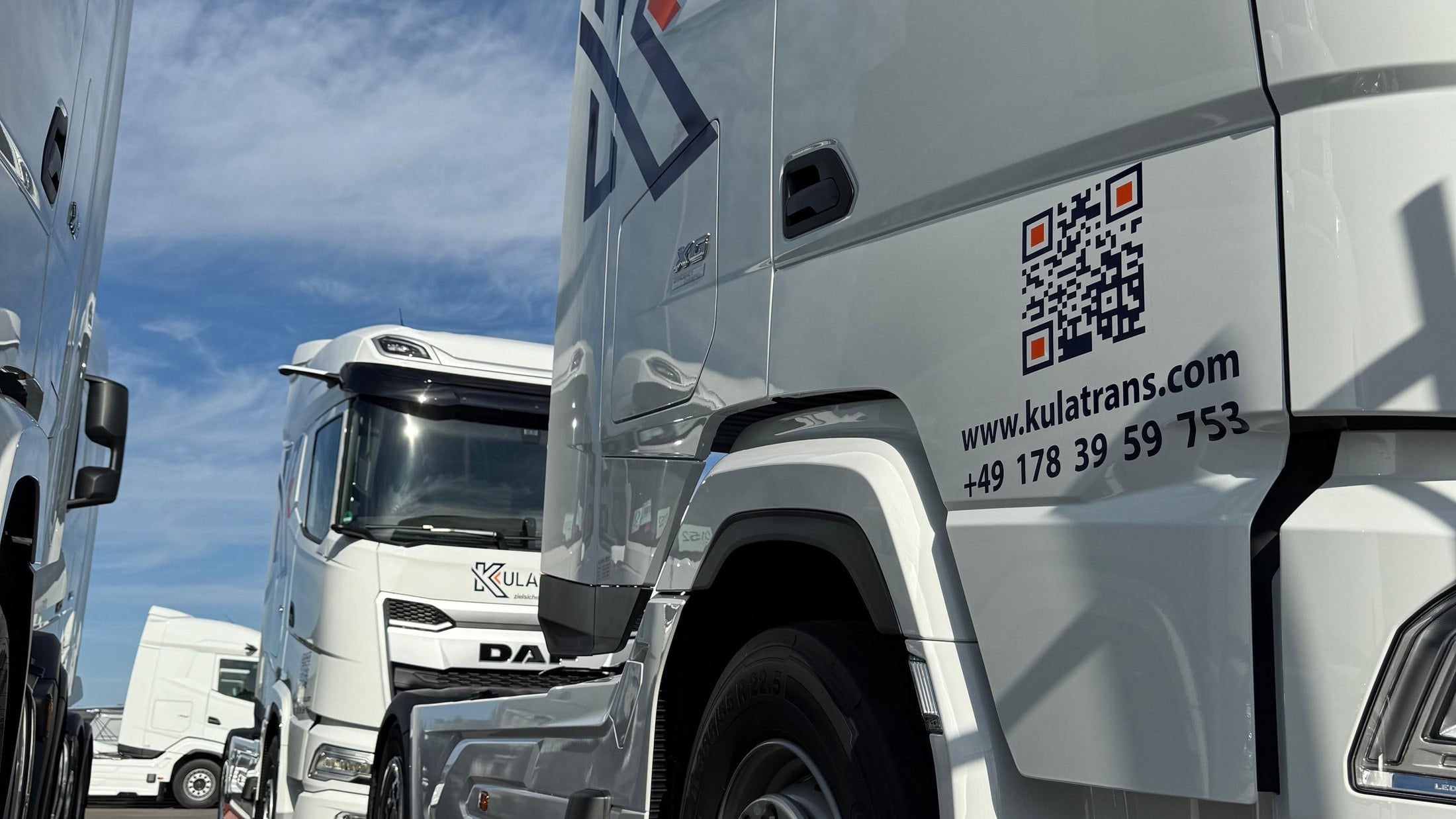 Mehrere weiße Sattelzugmaschinen von Kulatrans stehen dicht nebeneinander auf einem Hof, aufgenommen aus flachem Winkel gegen blauen Himmel. Auf dem vordersten Fahrerhaus stehen ein QR-Code, die Internetadresse und die Telefonnummer.